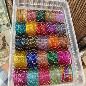 Puede incluir: Un recipiente de almacenamiento rectangular de plástico transparente lleno de filas de brazaletes finos con acabado metálico en varios colores, incluyendo oro, rosa, verde, azul, rojo, morado y naranja, con pequeños detalles dorados espaciados.