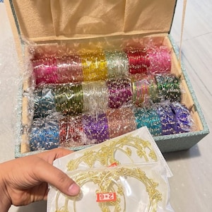May include: A collection of multi-colored glass bangles with gold-tone accents, organized in a light blue fabric-lined storage box. A separate plastic packet contains gold-tone wire bangles with a red label marked with the text 2*6.