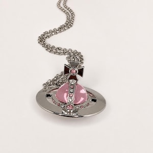 May include: A silver-toned necklace featuring a pendant shaped like a planetary orb. The orb has a translucent light pink spherical center, surrounded by a silver ring and a cross topper, all embellished with small clear and pink crystals.