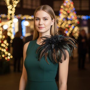 May include: A sleeveless dark green dress featuring a large, decorative shoulder accent composed of layered black and brown feathers with a central floral-shaped element. The garment has a simple, fitted bodice and a clean, rounded neckline.
