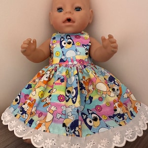 May include: A baby doll wearing a blue and orange dress with a rainbow and flower pattern. The dress has a white lace trim.