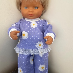 May include: A doll with short blonde hair and blue eyes wearing a two-piece pyjama set. The outfit consists of a long-sleeved top and matching trousers in a light purple fabric featuring a white polka dot pattern and a white daisy print.