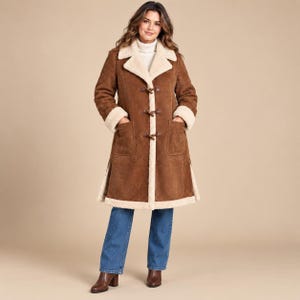 May include: A knee-length brown suede coat featuring a cream-colored faux shearling lining, a wide collar, and three toggle closures down the front. The coat includes two large patch pockets on the front and is worn over a cream turtleneck and blue jeans.