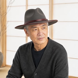 May include: A charcoal grey felt fedora hat featuring a structured crown and a wide brim. The hat is accented with a thin, dark brown leather band that encircles the base of the crown, providing a distinct contrast to the grey felt material.