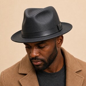Könnte beinhalten: Ein schwarzer Fedora-Hut mit einer texturierten, genarbten Oberfläche und einem glatten, einfarbigen schwarzen Band um die Krone. Der Hut hat eine eingedrückte Oberseite und eine strukturierte Krempe, was eine klassische Silhouette ergibt.