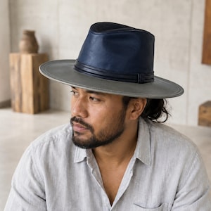May include: A fedora hat featuring a dark navy blue crown and a wide, flat grey brim. The hat is accented with a thin, dark navy blue band wrapped around the base of the crown, providing a distinct contrast between the blue top and grey brim.