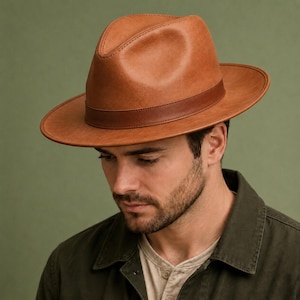 May include: A tan fedora hat with a structured crown and a wide brim. The hat features a smooth, matte finish and a matching tan band encircling the base of the crown. The design includes a pinched top and a clean, minimalist silhouette.