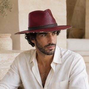 Könnte beinhalten: Ein dunkelroter Fedora-Hut mit einem dunkelbraunen Lederband um die Krone. Die Krempe ist breit und flach, wobei auf der Unterseite ein deutliches Leopardenmuster zu sehen ist. Der Hut ist mit sichtbaren hellen Nähten verarbeitet.