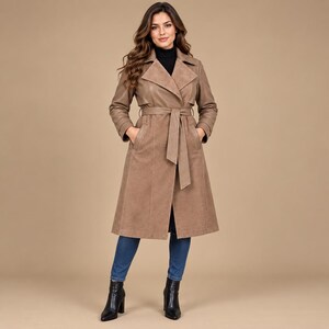 May include: A tan trench coat featuring a wide lapel, long sleeves with cuff straps, and a matching tie belt at the waist. The coat is worn over blue denim jeans and black ankle boots, creating a structured silhouette with a smooth finish.