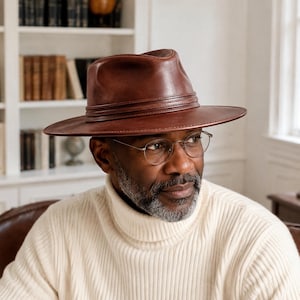 Puede incluir: Un sombrero fedora de cuero marrón con una copa estructurada y ala ancha, que presenta una banda de cuero a juego envuelta alrededor de la base de la copa. El sombrero tiene un acabado liso y pulido con una silueta clásica de parte superior pellizcada.