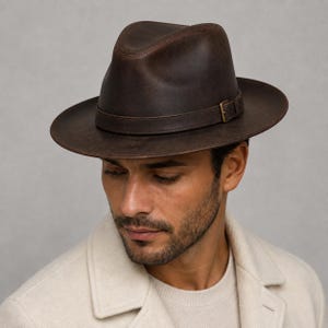Op de afbeelding: Een donkerbruine fedora hoed met een ingedeukte kroon en een brede rand. De hoed is voorzien van een bijpassende donkerbruine leren band rond de basis, geaccentueerd met een kleine, rechthoekige, messingkleurige metalen gesp aan de zijkant.