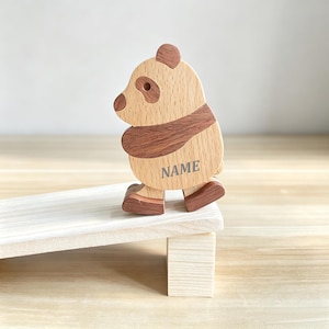 Pode incluir: Uma figura de panda de madeira feita com peças de madeira clara e castanho-escura. O panda apresenta um corpo arredondado, orelhas pequenas e uma mancha escura no olho. A palavra NAME está impressa em letras maiúsculas cinzentas no corpo.