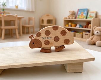 May include: A wooden ladybird toy featuring a light and dark brown two-tone finish with circular spots and the word NAME printed on the side. The toy has two small antennae and is mounted on two small wooden rockers for movement.