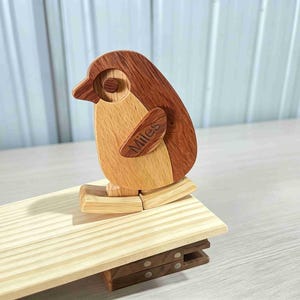 May include: A wooden penguin figurine crafted from two-toned light and dark wood, featuring the name Miles engraved on its wing. The penguin is mounted on a light-coloured wooden plank base, which is attached to a dark wooden clip mechanism.