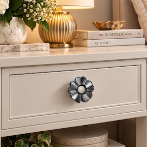 May include: A flower-shaped drawer knob with a dark, metallic finish and a smooth, white circular centre. The knob is attached to the front of a light-coloured wooden nightstand drawer, featuring a simple, clean design with six rounded petals.