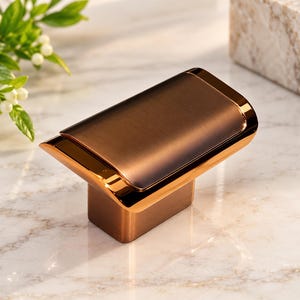 May include: A rectangular cabinet knob featuring a brushed bronze finish on the top surface and polished copper-toned edges. The hardware has a wide, slightly curved top section that tapers down to a smaller, solid rectangular base for mounting.