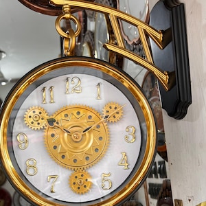 Op de afbeelding: Een dubbelzijdige wandklok met een goudkleurige ronde rand en een witte wijzerplaat met verhoogde gouden cijfers. Het midden toont zichtbare gouden mechanische tandwielen en zwarte wijzers. Gemonteerd op een zwarte beugel met gouden accenten.