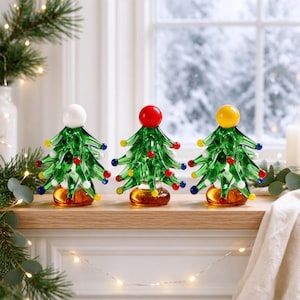 May include: Three glass Christmas tree figurines with green branches and multicoloured glass bead ornaments. Each tree features a distinct solid-coloured sphere at the top: one white, one red, and one yellow, all resting on amber-coloured bases.
