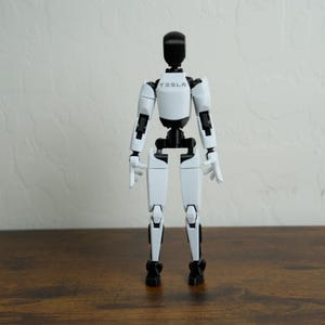 May include: A white and black humanoid robot action figure with a smooth finish. The chest area features the word TESLA printed in black. The figure has articulated joints at the shoulders, elbows, hips, and knees, standing in a neutral pose.