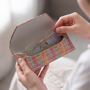 Könnte beinhalten: Eine Brille mit dünnem Goldmetallrahmen in sechseckiger Form, die in einem Hartschalenetui liegt, das ein lebendiges, mehrfarbig gewebtes Außenmaterial mit strukturiertem Muster und ein glattes, hellgraues Innenfutter aufweist.