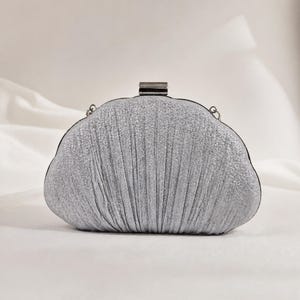 May include: A silver glittery clutch bag with a rounded, shell-like shape and vertical pleated fabric texture. The bag features a metallic rectangular clasp closure at the top centre and small metal loops on the sides for an optional strap attachment.