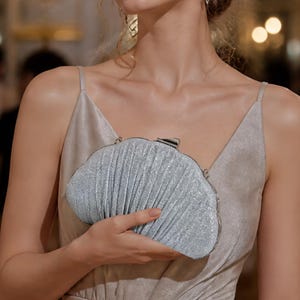 May include: A silver glittery clutch bag with a pleated texture and a metallic frame closure. The handbag features a curved, shell-like silhouette with a shimmering finish, designed to be held in the hand for formal occasions.
