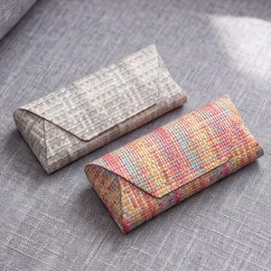 May include: Two rectangular spectacle cases with triangular fold-over flaps. One case features a neutral grey woven fabric texture, while the other displays a vibrant, multicoloured woven pattern in shades of pink, yellow, and light blue.