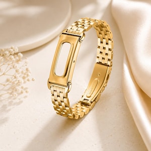 May include: A gold-toned stainless steel replacement watch band featuring a polished rectangular frame for a fitness tracker. The band consists of interconnected metallic links and includes a secure clasp mechanism on the underside of the strap.