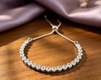 Pode incluir: Uma pulseira de ténis em tom prateado com uma fila contínua de pedras transparentes de corte quadrado. A pulseira inclui uma corrente delicada com um mecanismo de fecho de conta deslizante para ajustar a diferentes medidas de pulso.
