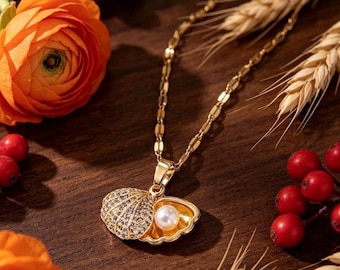 Op de afbeelding: Een goudkleurige ketting met een hanger in de vorm van een geopende schelp. De buitenkant van de schelp is bezet met kleine doorzichtige steentjes, terwijl de binnenkant een enkele witte, bolvormige parel bevat. De hanger hangt aan een fijne ketting.