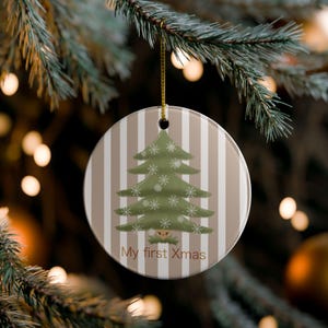 Può includere: Un ornamento rotondo in ceramica con uno sfondo a righe verticali beige e bianche. Un albero di Natale verde con motivi a fiocchi di neve bianchi è centrato. Il testo "My first Xmas" è stampato in marrone nella parte inferiore.