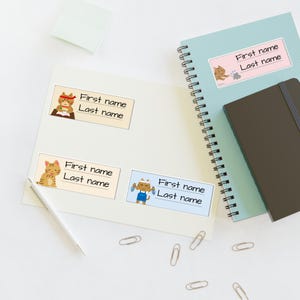 Pode incluir: Quatro etiquetas adesivas retangulares com ilustrações de gatos de desenho animado. Cada etiqueta inclui o texto impresso First name e Last name com uma linha em branco para escrever. Os gatos são retratados em várias atividades, incluindo estudar e brincar.