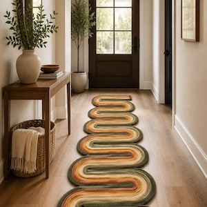 May include: A long, narrow runner rug featuring a continuous, winding serpentine pattern in shades of olive green, burnt orange, cream, and tan. The rug is laid out on a light-colored wooden floor, extending from the foreground toward a dark wooden door.