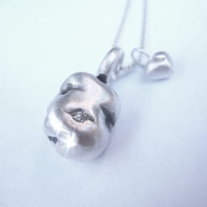 May include: A silver necklace with a textured pendant featuring a small diamond. The pendant is shaped like a face with a single eye. The necklace also has a small heart charm.