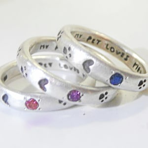 May include: Three silver rings with paw prints and hearts engraved on them. The rings are stacked on top of each other and have small colorful gemstones embedded in them. The text on the rings reads "My Pet Loves Me".