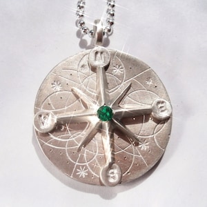 May include: A silver compass pendant with a green gemstone in the center. The compass has a star-shaped design with four points and is surrounded by a circle of smaller stars.