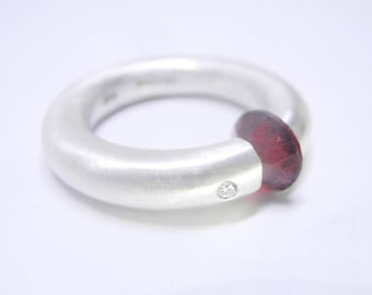 Könnte beinhalten: Ein silberner Ring mit gebürsteter Oberfläche und einem kleinen Diamanten, der in die Ringschiene eingefasst ist. Ein dunkelroter Edelstein ist in den Ring eingefasst und bedeckt die Ringschiene teilweise.
