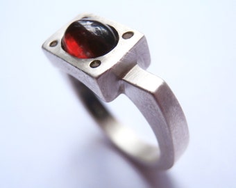 May include: A silver ring with a square setting featuring a red and black gemstone.