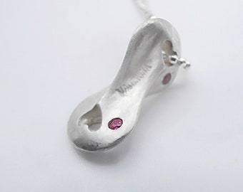 May include: A silver pendant with a unique, abstract shape. The pendant has a matte finish and features three small, round, pink gemstones. The word "VALENCIA" is engraved on the side. The pendant is on a silver chain.
