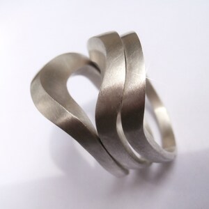 May include: Three silver rings, each with a unique wave-like design, are stacked on top of each other. The rings are made of a smooth, polished metal and have a contemporary style.
