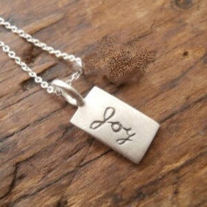 May include: A silver rectangular pendant necklace with the word "Joy" engraved in cursive. The pendant hangs from a delicate silver chain. The necklace rests on a textured, dark wooden surface.