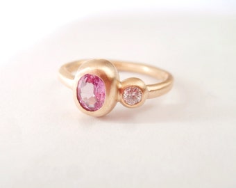 May include: A gold ring featuring a large oval pink gemstone and a smaller round clear gemstone. The ring has a simple band and a modern design, perfect for everyday wear.