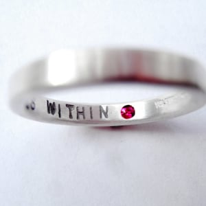 May include: A silver ring with the word "WITHIN" engraved on the band and a small red gemstone set in the center.