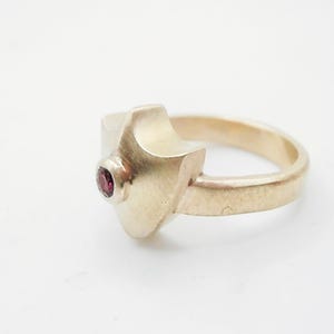 May include: A gold ring with a geometric design and a small red gemstone.
