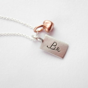 May include: A silver necklace with a silver tag charm that says "Be" and a small rose gold heart charm.
