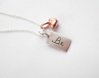 Könnte beinhalten: Eine silberne Halskette mit einem silbernen Anhänger in Form eines Schildchens, auf dem "Be" steht, und einem kleinen roségoldenen Herzanhänger.
