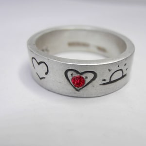 以下が含まれることがあります： 赤いハートと太陽のデザインが施された銀の指輪。指輪にはハートの中心に小さな赤い宝石が埋め込まれています。