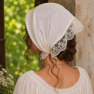 May include: An off-white triangular headscarf made of a textured fabric, featuring a delicate white lace trim along the edges and a small pom-pom fringe detail along the front border, tied securely at the nape of the neck.