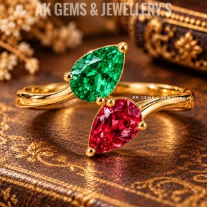 May include: A gold bypass ring featuring two pear-cut gemstones, one vibrant green and one deep red, set in a polished gold band. The text AK GEMS & JEWELLERY'S is visible at the top and center of the image. The ring has a sleek, open design.