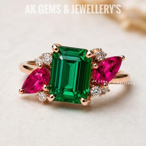 Könnte beinhalten: Ein Ring aus Roségold mit einem großen smaragdgeschliffenen grünen Mittelstein, flankiert von zwei birnenförmigen rosa Steinen und vier kleinen runden weißen Steinen. Der Text AK GEMS & JEWELLERY'S ist oben und mittig am Ring zu sehen.
